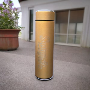 A tumbler standing on a concrete surface outside a house. The tumbler features the name Summer written. In the background, there is a window with closed blinds and a potted plant with purple flowers.