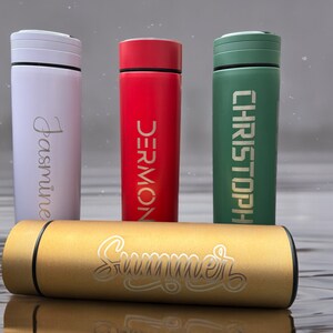 Four thermos bottles stand on a wet surface. From left to right: Jasmine (white), DERMONT (red), CHRISTOPHER (green). A gold bottle labeled Summer lies horizontally in the foreground.