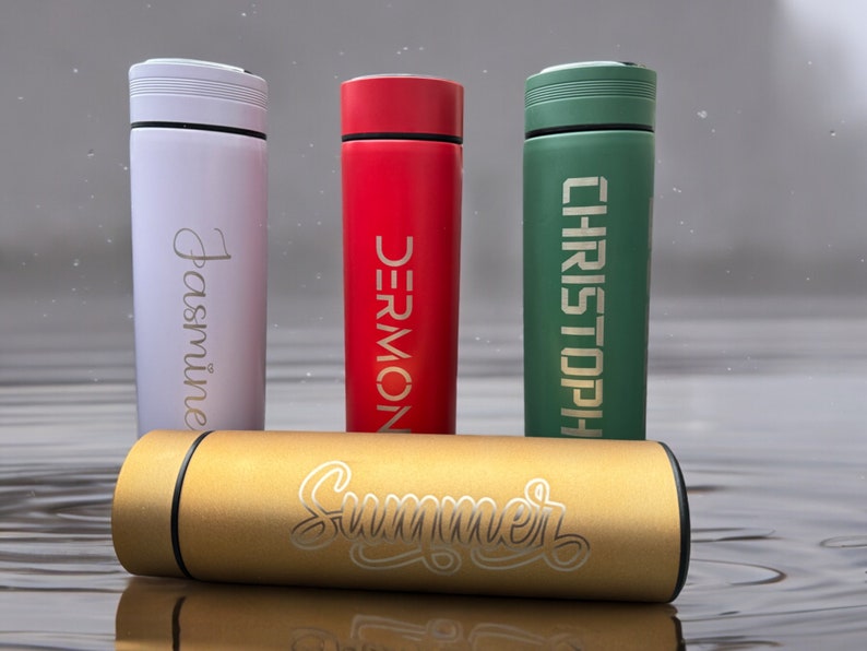 Four thermos bottles stand on a wet surface. From left to right: Jasmine (white), DERMONT (red), CHRISTOPHER (green). A gold bottle labeled Summer lies horizontally in the foreground.