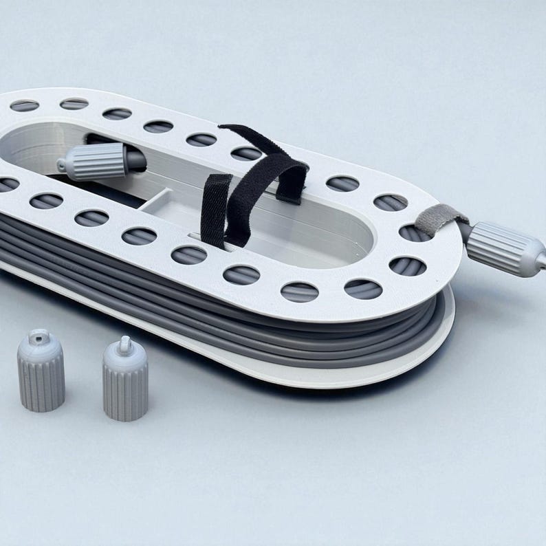 May include: A gray and white cable organizer with a series of oval holes and a black strap. Two gray caps are in the foreground. The organizer is designed to hold and manage cables, keeping them neat and tidy.