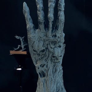May include: A detailed, gray sculpture of a hand with long, pointed fingers. The hand is textured with a rough, weathered appearance, and features carved skull-like faces. A small, dark creature sits on a shelf in the background, adding to the eerie atmosphere.
