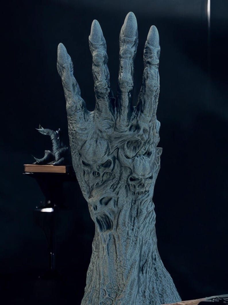 May include: A detailed, gray sculpture of a hand with long, pointed fingers. The hand is textured with a rough, weathered appearance, and features carved skull-like faces. A small, dark creature sits on a shelf in the background, adding to the eerie atmosphere.