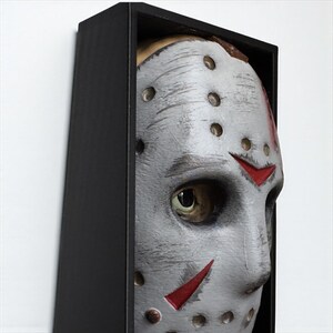 May include: A gray and white hockey mask with red accents, displayed in a black shadow box. The mask has visible eye holes and is a close-up shot. The mask is a prop from a horror movie.