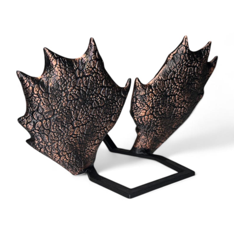 May include: A decorative metal sculpture of two stylized wings with a textured, copper-colored finish. The wings are mounted on a black metal base.