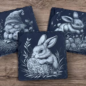 Engraved slate coaster featuring a detailed illustration of a bunny resting on a decorative Easter egg, surrounded by grass and leaves. The coaster has a natural, rough-edged finish and is set on a wooden background