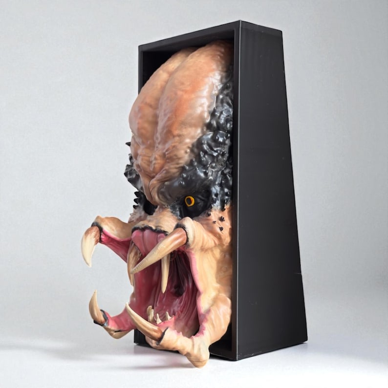 May include: A detailed sculpture of a Predator alien head, featuring a textured, tan and black skin tone, sharp teeth, and yellow eyes. The sculpture is partially encased in a black rectangular frame, creating a 3D effect.