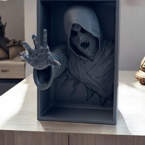 May include: A 3D printed sculpture of a hooded figure with a menacing expression reaching out from a gray box. The figure is made of a dark gray material and appears to be emerging from the box.