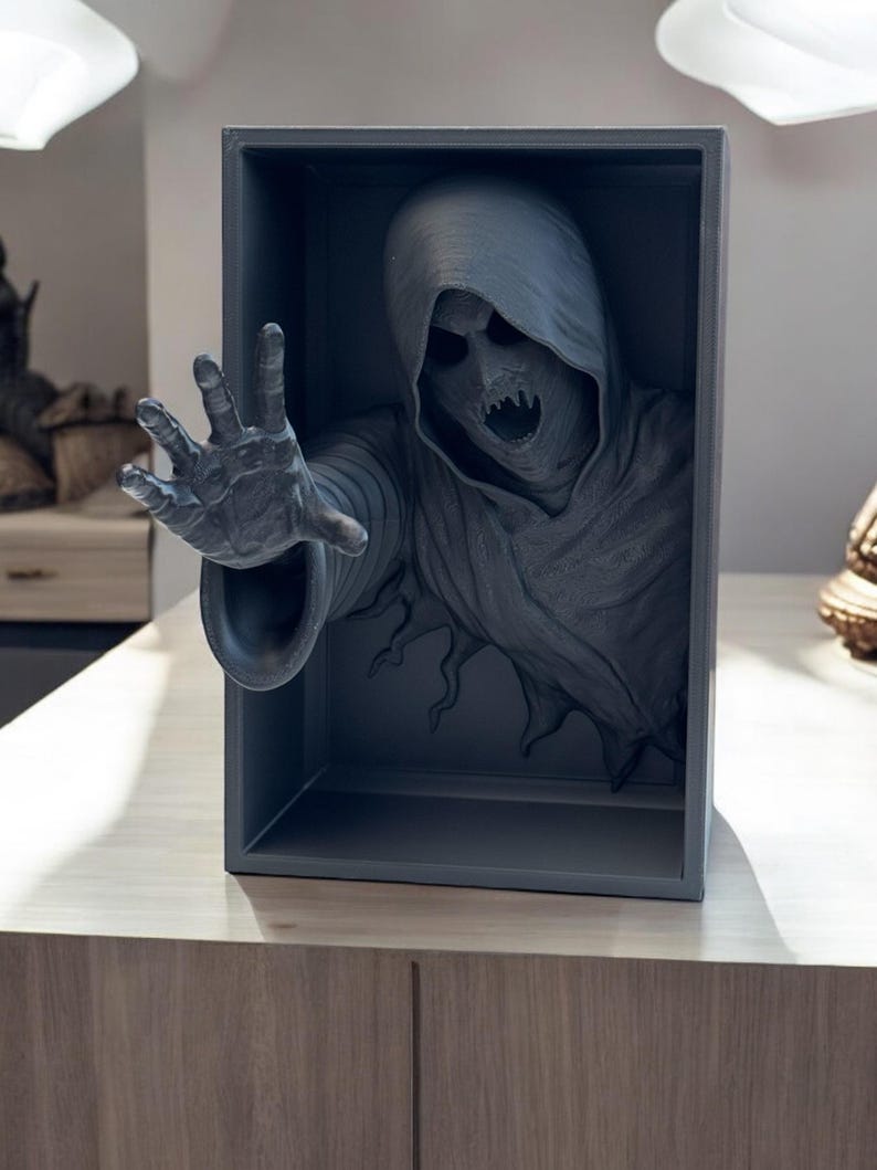 May include: A 3D printed sculpture of a hooded figure with a menacing expression reaching out from a gray box. The figure is made of a dark gray material and appears to be emerging from the box.