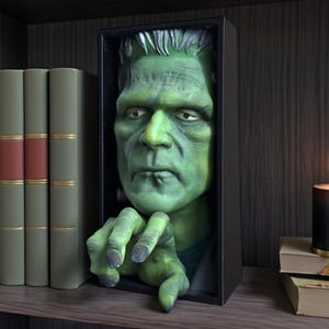 May include: A green Frankenstein bust sculpture in a black box frame. The monster's face and hand extend from the frame, creating a 3D effect. The sculpture is displayed on a shelf with books and a candle.