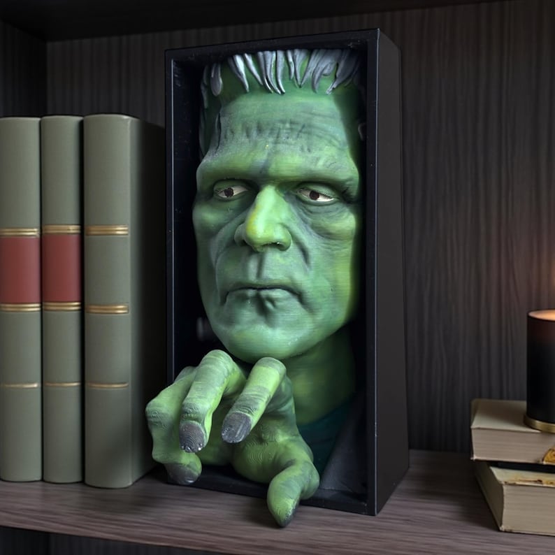 May include: A green Frankenstein bust sculpture in a black box frame. The monster's face and hand extend from the frame, creating a 3D effect. The sculpture is displayed on a shelf with books and a candle.