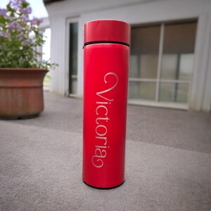 A tumbler standing on a concrete surface outside a house. The tumbler features the name Victoria written. In the background, there is a window with closed blinds and a potted plant with purple flowers.