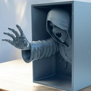 May include: A 3D printed sculpture of a hooded figure reaching out from a box. The figure is gray and has a skeletal face.