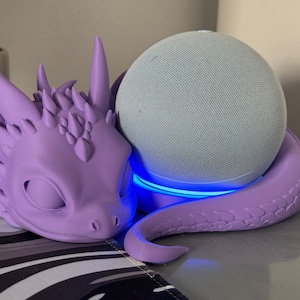 Pode incluir: Um suporte em forma de drag&atilde;o roxo com um brilho azul claro ao redor da base, segurando um alto-falante inteligente cinza e branco.