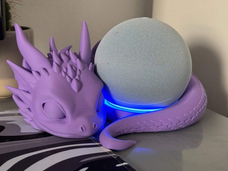 Pode incluir: Um suporte em forma de drag&atilde;o roxo com um brilho azul claro ao redor da base, segurando um alto-falante inteligente cinza e branco.