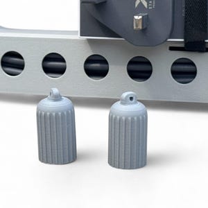 May include: Two light gray, cylindrical caps with vertical ridges and a small loop at the top. The caps are in front of a light gray, rectangular structure with circular openings and a black cable.