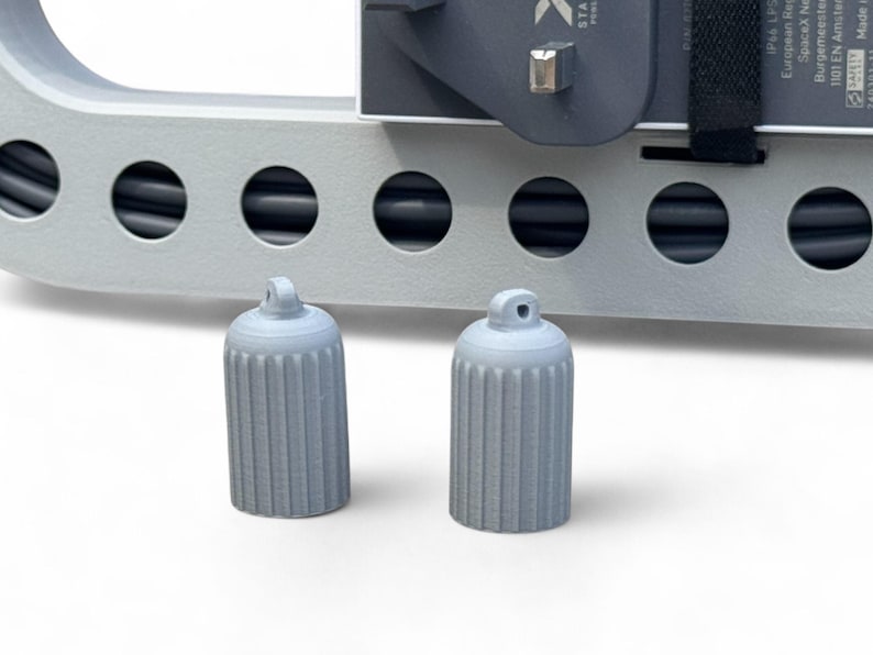 May include: Two light gray, cylindrical caps with vertical ridges and a small loop at the top. The caps are in front of a light gray, rectangular structure with circular openings and a black cable.
