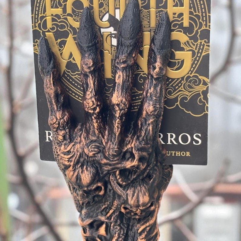 May include: A bronze-colored, detailed hand-shaped book holder with black fingertips. The book holder is holding a book with the title "Fourth Wing" and the author's name. The background is blurred, with tree branches visible.