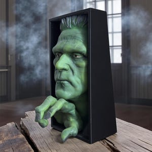 May include: A green Frankenstein monster head and hand emerging from a black rectangular box. The monster has green skin, black hair, and a serious expression. The hand is reaching out. The box is on a wooden surface.