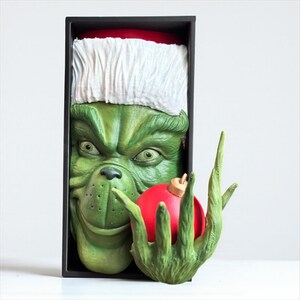 May include: A green Grinch sculpture wearing a Santa hat, holding a red Christmas ornament. The Grinch's face is detailed with a mischievous grin. The sculpture is in a black box, perfect for holiday decor.