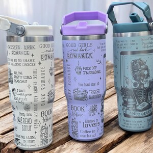 May include: Three stainless steel tumblers with different designs. The first tumbler is white with black text that says "Twisted, Dark, Romance" and other related phrases. The second tumbler is purple with black text that says "Good Girls Read Dark Romance" and other related phrases. The third tumbler is blue with a black and white illustration of a skeleton with a book. The text on the tumbler says "Spread Them  Thick  Baby  Like  Butter  On  A  Scone" and "Smut Queen".