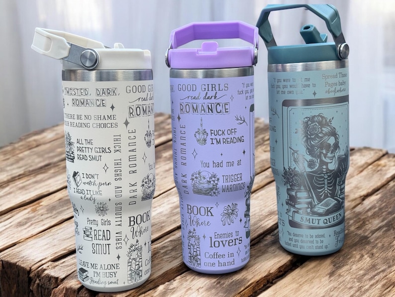 May include: Three stainless steel tumblers with different designs. The first tumbler is white with black text that says "Twisted, Dark, Romance" and other related phrases. The second tumbler is purple with black text that says "Good Girls Read Dark Romance" and other related phrases. The third tumbler is blue with a black and white illustration of a skeleton with a book. The text on the tumbler says "Spread Them  Thick  Baby  Like  Butter  On  A  Scone" and "Smut Queen".