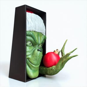May include: A green Grinch figure wearing a Santa hat, emerging from a black box, holding a red Christmas ornament. The Grinch's face is detailed with a mischievous grin and yellow eyes. The ornament has a gold cap.