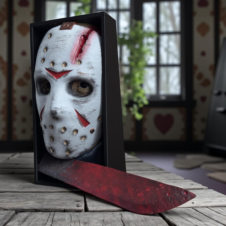 May include: A white hockey mask with red accents and a slash mark, displayed in a black box with a blood-stained machete. The mask has eye and mouth openings and is a horror movie prop.
