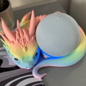 Pode incluir: Uma figura de drag&atilde;o com cores de arco-&iacute;ris segurando um alto-falante inteligente azul claro. O alto-falante tem uma luz azul ao redor da base.