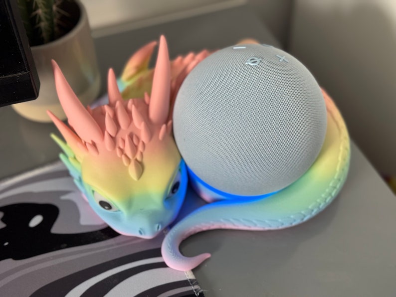 Pode incluir: Uma figura de drag&atilde;o com cores de arco-&iacute;ris segurando um alto-falante inteligente azul claro. O alto-falante tem uma luz azul ao redor da base.