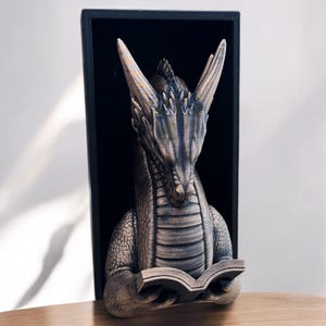 May include: A gray dragon bookend with a book in its claws. The dragon is in a black frame and is looking down at the book.
