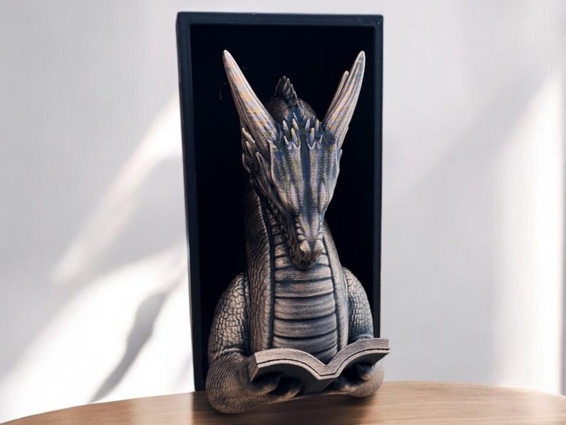May include: A gray dragon bookend with a book in its claws. The dragon is in a black frame and is looking down at the book.