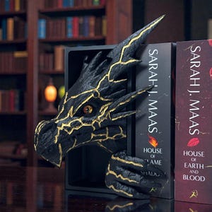 以下が含まれることがあります： 黒いドラゴンブックエンド、金色のアクセント付き。黒い箱から現れています。ドラゴンの頭と爪が見え、本には「SARAH J. MAAS」の文字と本のタイトルが書かれています。