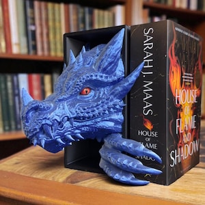 May include: A blue dragon bookend with a detailed, textured design. The dragon's head and claw emerge from a black bookend, with a red eye visible. The bookend is next to a book titled "House of Flame and Shadow."