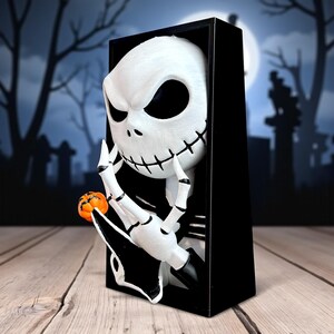 May include: A white Jack Skellington figure with black eyes and stitched mouth, holding a small orange pumpkin. The figure is emerging from a black rectangular frame, with a spooky graveyard scene in the background. A Halloween-themed decoration.