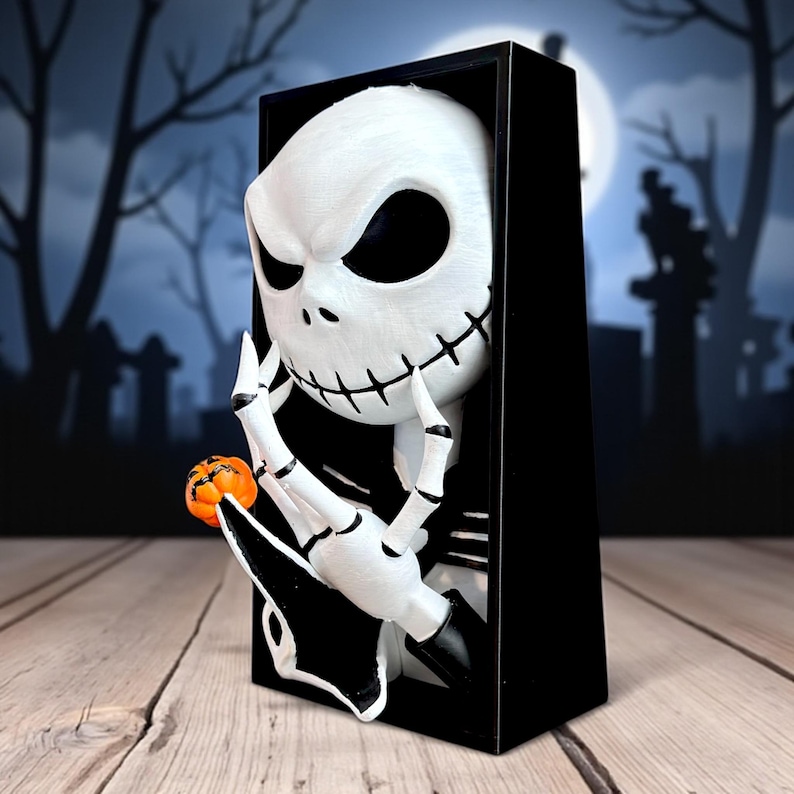 May include: A white Jack Skellington figure with black eyes and stitched mouth, holding a small orange pumpkin. The figure is emerging from a black rectangular frame, with a spooky graveyard scene in the background. A Halloween-themed decoration.