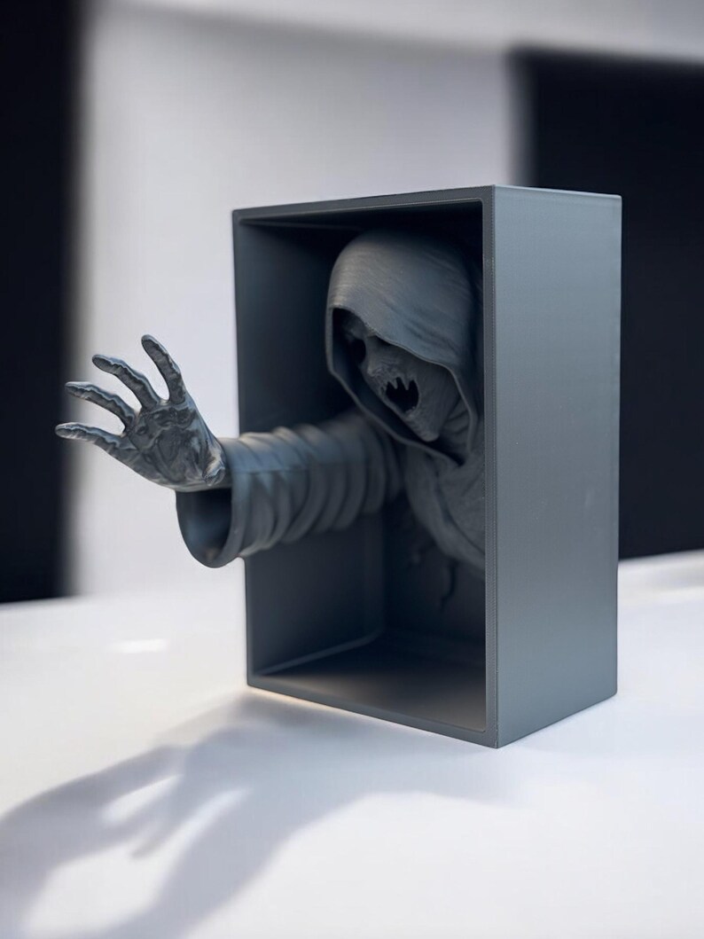 May include: A gray 3D printed sculpture of a hooded figure with a skeletal face reaching out from a gray box.
