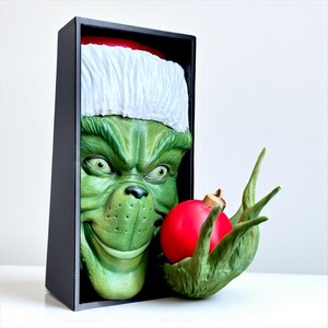 May include: A green Grinch figure wearing a Santa hat, holding a red Christmas ornament. The Grinch's face is framed in a black box. The ornament is held in a green hand with long fingers. The Grinch has a mischievous grin.