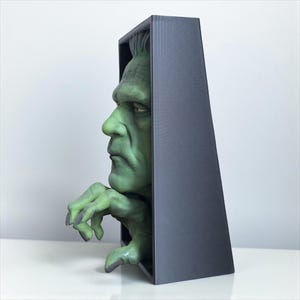 May include: A green Frankenstein monster bust emerges from a dark gray rectangular frame. The monster's face and hand are visible, with detailed features and a textured appearance. The sculpture is set against a plain white background.