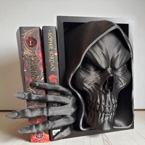 May include: A black skull-shaped bookend with a skeletal hand holding two books. The bookend has a detailed, textured finish. The books are black with red and gold accents. The bookend is designed to hold books upright.