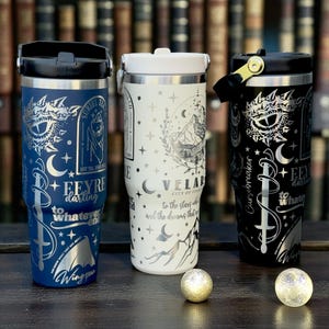 May include: Three insulated travel mugs in black, white, and blue. Each mug has silver designs, including text, celestial elements, and a dragon. The mugs are on a wooden surface, with two small decorative spheres.
