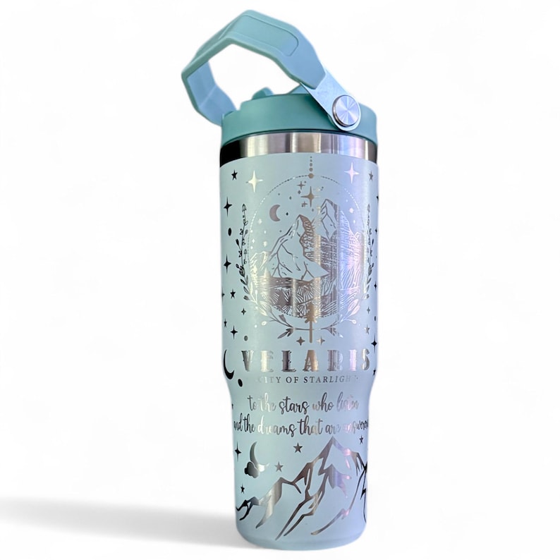 May include: A light blue insulated tumbler with a silver mountain and star design. The tumbler has a matching light blue handle and lid. Text on the tumbler reads "VELARIS CITY OF STARLIGHTS to the stars who listen and the dreams that are answered."