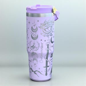 May include: A lilac insulated travel mug with a matching lid and handle. The mug features celestial and fantasy-themed designs, including moons, stars, and a sword. The text "Cursebreaker" is printed on the side.