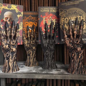 May include: Four decorative hand sculptures with a gnarled, textured appearance, each in a different color: bronze, dark brown, black, and a lighter brown. The hands are positioned in front of a bookshelf with book titles visible.