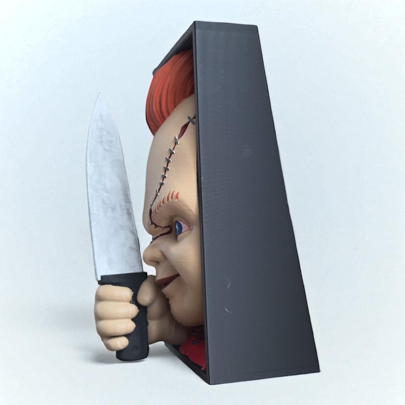 Puede incluir: Una cabeza de mu&ntilde;eco Chucky impresa en 3D con cabello rojo y detalles faciales cosidos, sosteniendo un cuchillo plateado. La mu&ntilde;eca est&aacute; parcialmente enmarcada por una estructura rectangular negra.