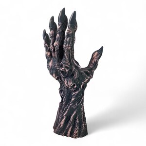 May include: A dark, textured sculpture of a hand with long, sharp fingernails. The hand is a mix of dark brown and bronze colors, with a detailed, almost skeletal appearance. The fingers are spread, and the palm is open.