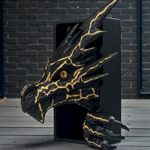 以下が含まれることがあります： 黒いドラゴンの頭の彫刻で、金色のアクセントが黒い長方形のフレームから現れています。 ドラゴンは詳細な質感、金色の目、鋭い爪を持っています。 彫刻は暗いレンガの壁を背景にしています。