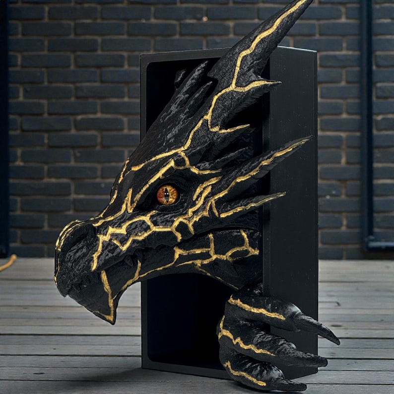 以下が含まれることがあります： 黒いドラゴンの頭の彫刻で、金色のアクセントが黒い長方形のフレームから現れています。 ドラゴンは詳細な質感、金色の目、鋭い爪を持っています。 彫刻は暗いレンガの壁を背景にしています。