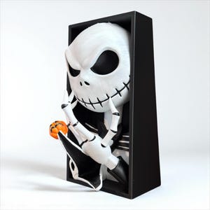 May include: A white Jack Skellington figure with black eyes and stitched mouth, holding a small orange pumpkin. The figure is set in a black rectangular frame, creating a 3D effect. A Halloween-themed decorative item.