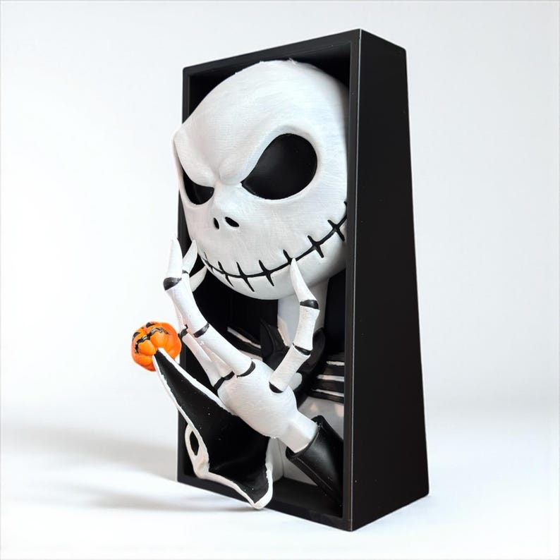 May include: A white Jack Skellington figure with black eyes and stitched mouth, holding a small orange pumpkin. The figure is set in a black rectangular frame, creating a 3D effect. A Halloween-themed decorative item.