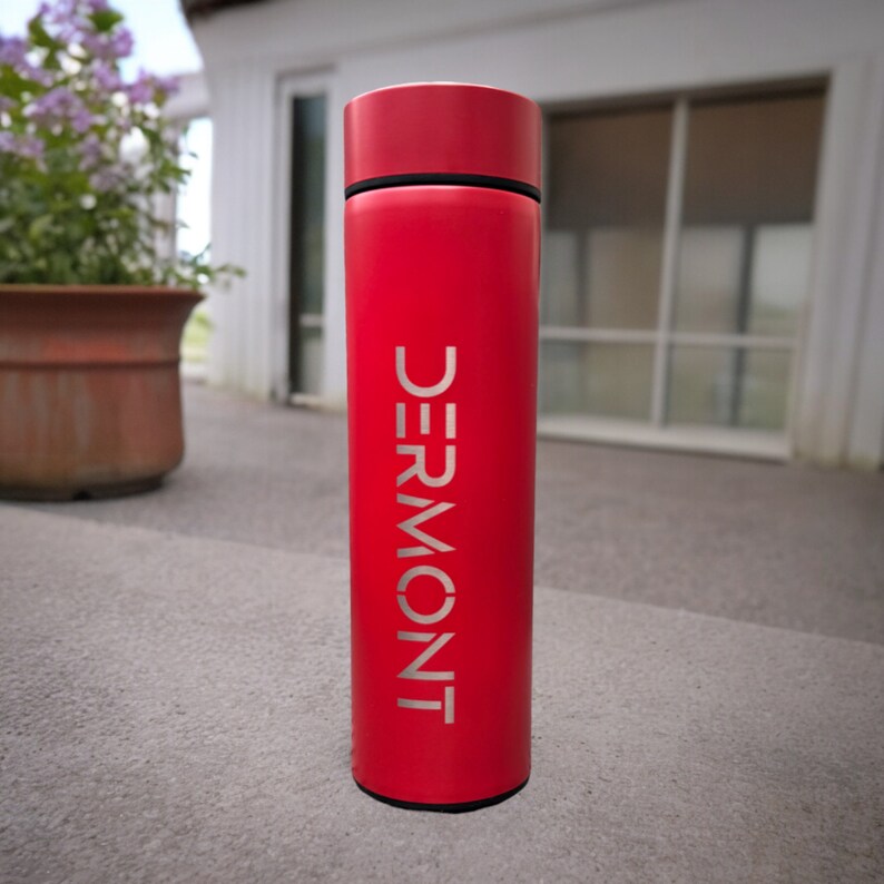 A tumbler standing on a concrete surface outside a house. The tumbler features the name Dermont written. In the background, there is a window with closed blinds and a potted plant with purple flowers.